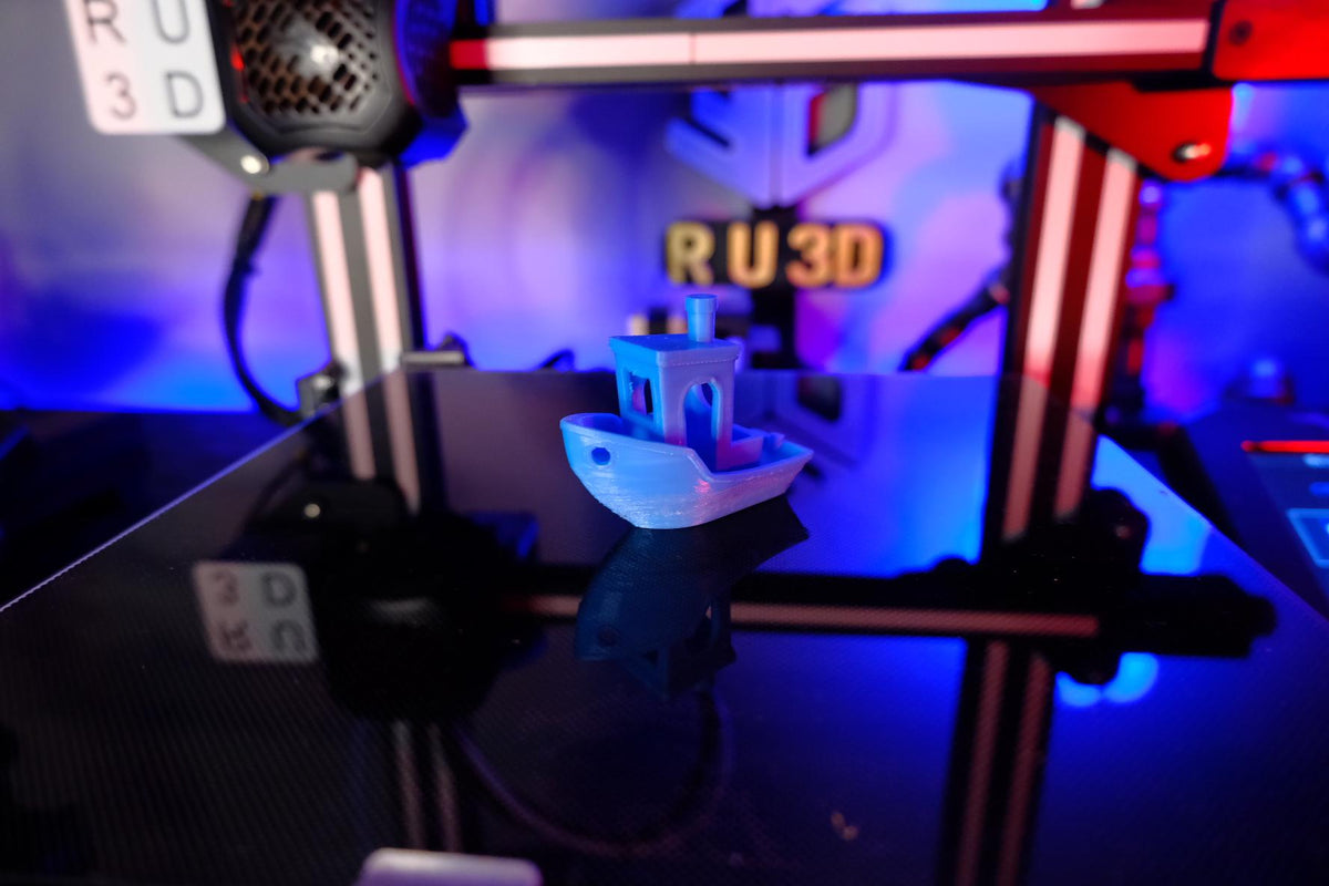 A 3D printed 3D Benchy boat sitting on a 3D printer with an RU3D logo in the background.