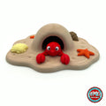Cute lobster keychain, 3D printed by RU3D – peeking out of beach cave playset. Bright red with sandy beach cave playset.