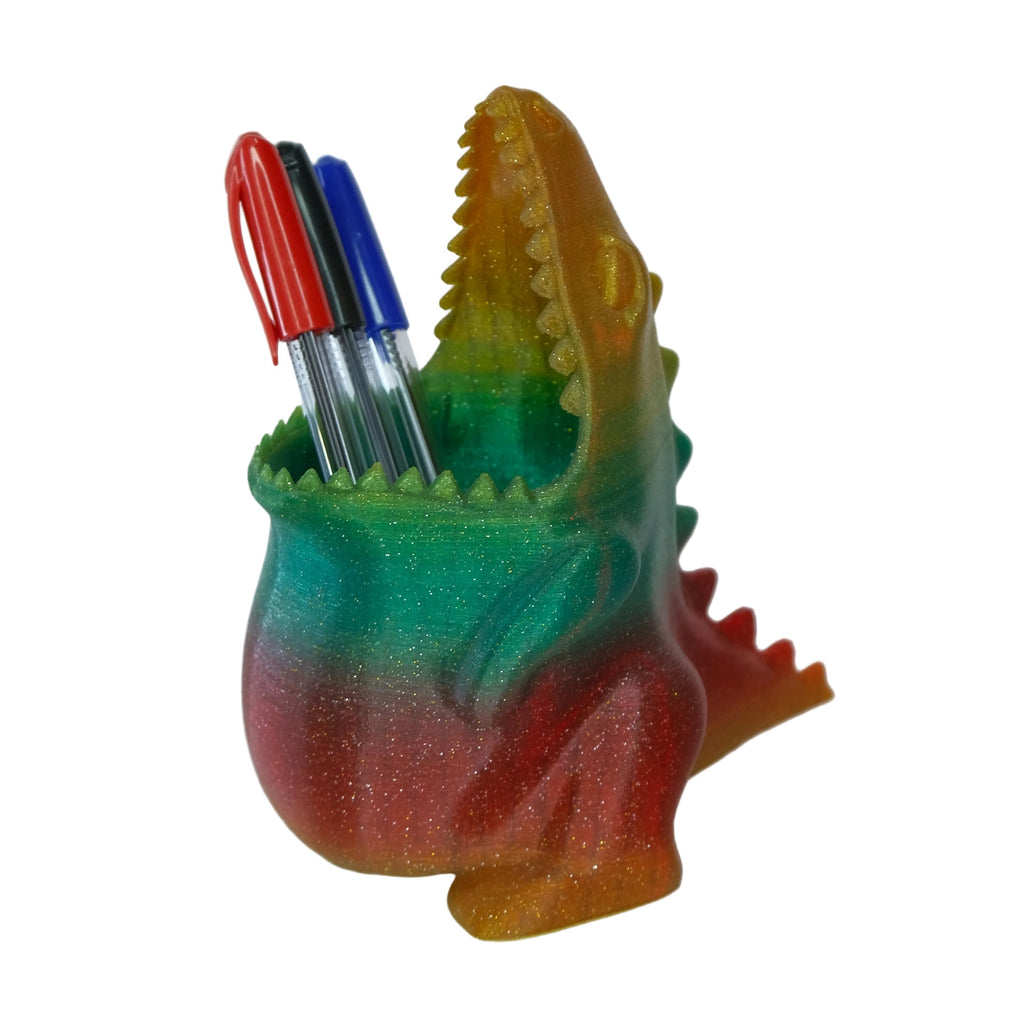 A novelty pen holder shaped like a crocodile, displaying a gradient of green, red, and yellow colours and holding several pens in its mouth.